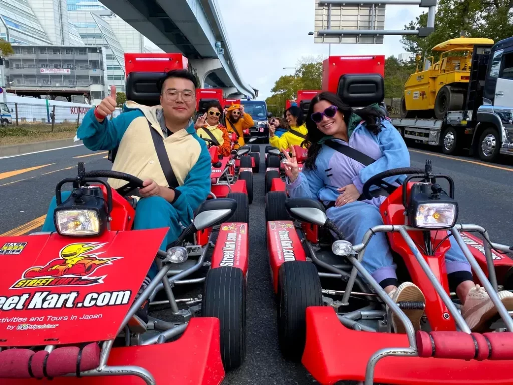 Two people in colorful onesies sit in red go-karts on a city street, smiling and giving thumbs up, part of a street kart tour.