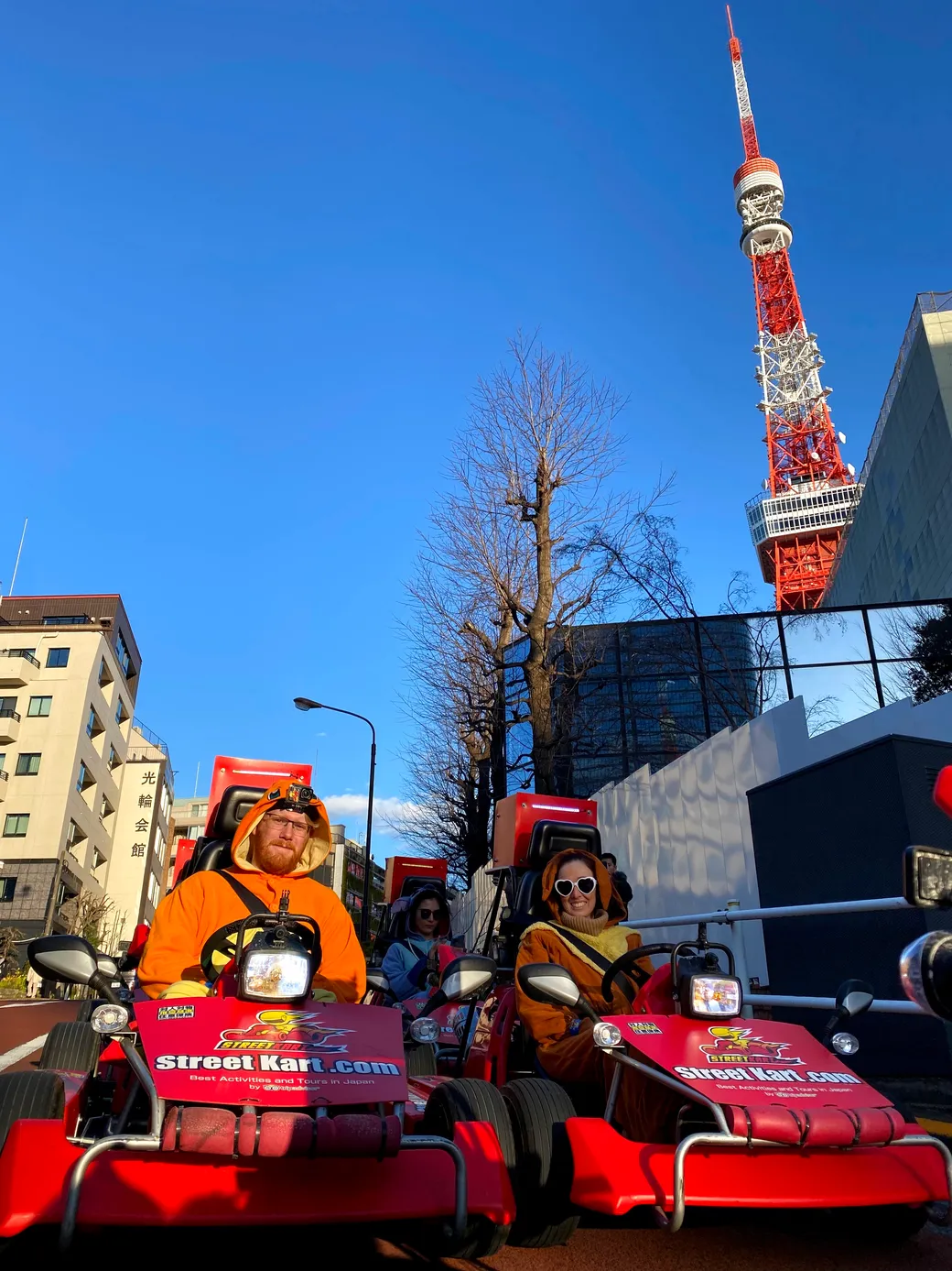 People in red Street Kart vehicles riding on a city street with a blue sky; Tokyo Tower visible in the background.
