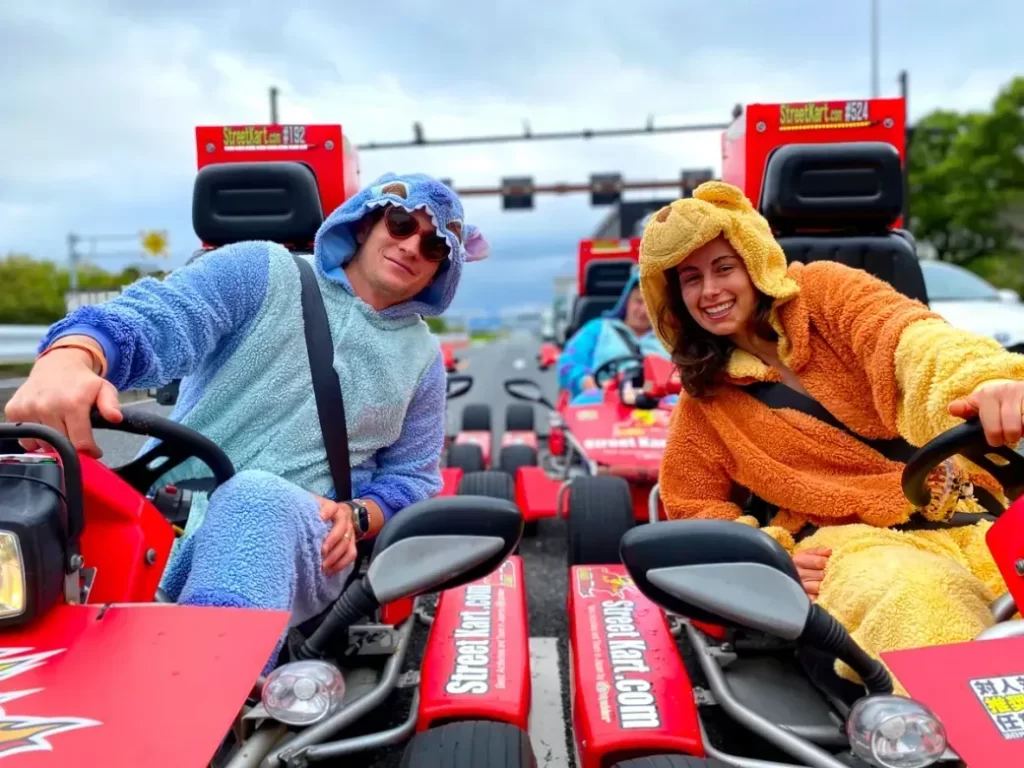 IMG_6138 - StreetKart Two people in blue and orange fluffy animal onesies drive red go-karts, smiling at the camera on a road course.